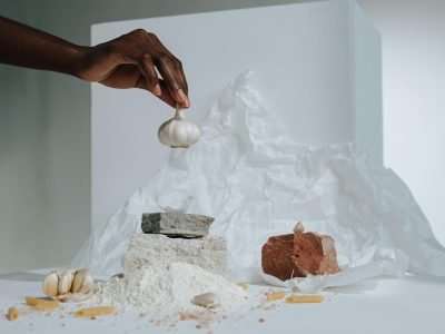 Artistic composition featuring a hand arranging garlic with pasta ingredients in a studio setting.