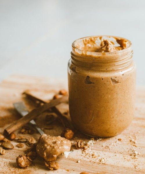 A jar of creamy peanut butter with scattered peanuts and spoon on a wooden surface.